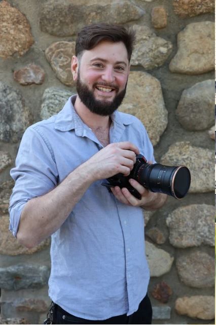 Man smiling while holding a camera in front of a stone wall
