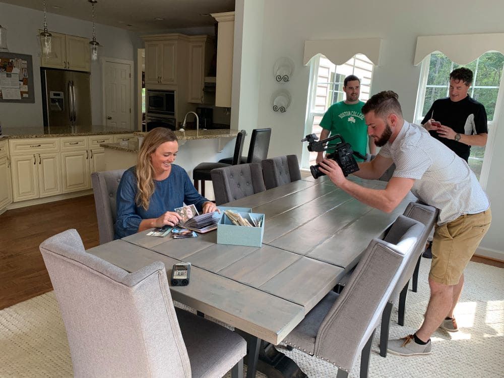 People in a bright dining room around a long table, with one person filming or photographing another.