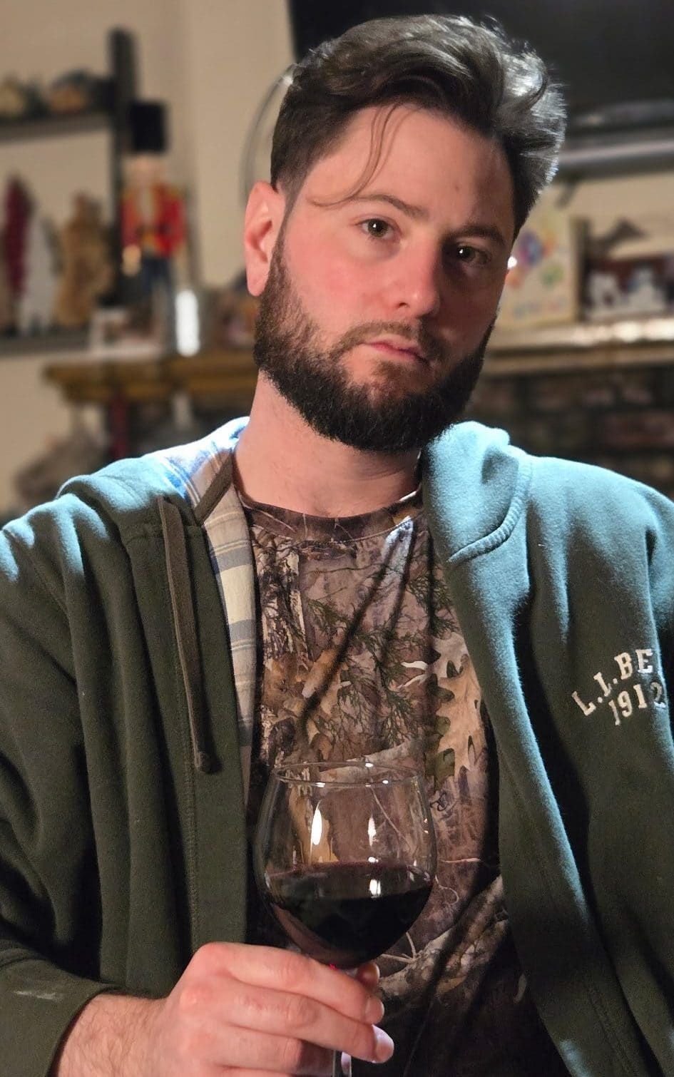 Person in a green hoodie and patterned shirt holding a glass of red wine, seated indoors with a thoughtful expression