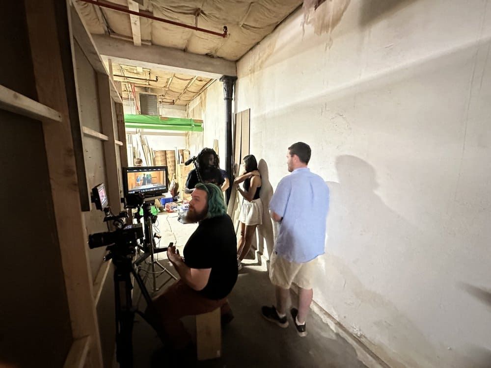 Several people work in a narrow hallway with cameras and a monitor, suggesting a film or photo shoot.