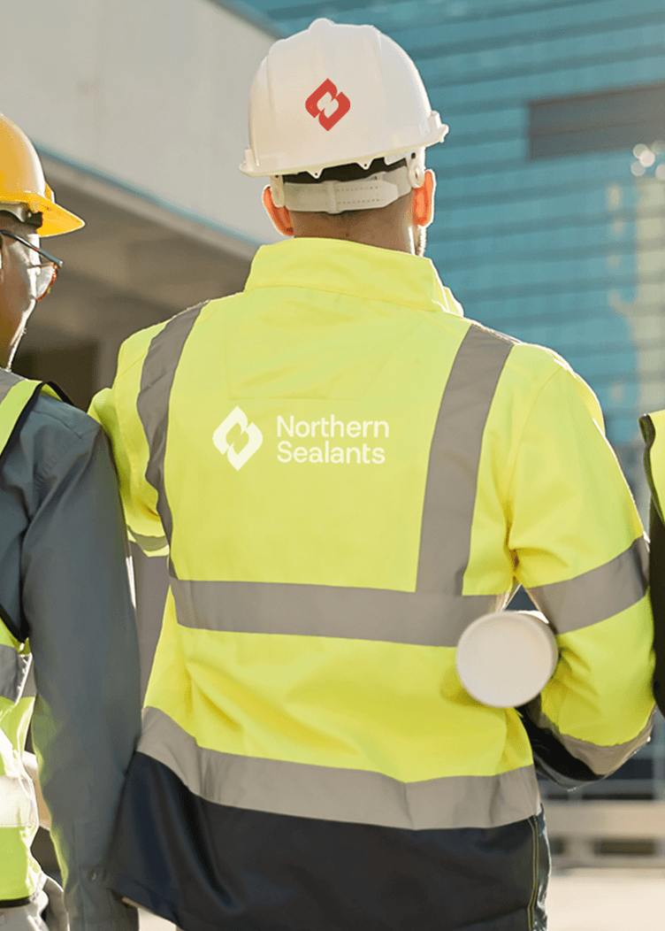 Two construction workers in hi-vis vests and hard hats at a building site, one with a Northern Sealants logo.