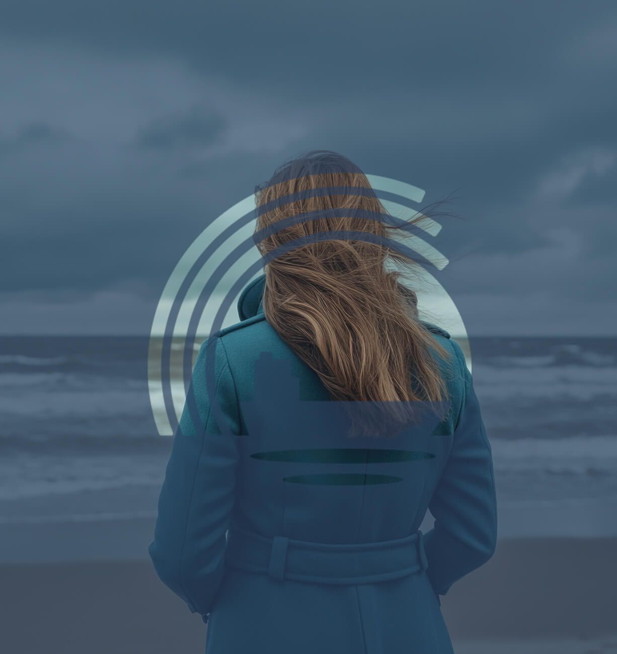 Person in a blue coat standing on a cloudy beach, facing the ocean
