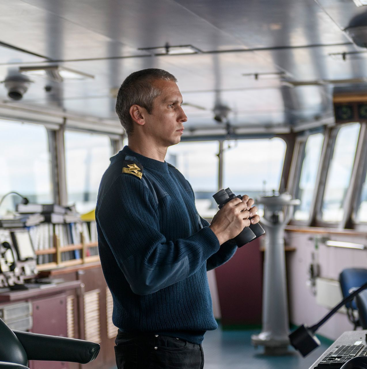 Man in a navy sweater standing on a ship’s bridge, looking ahead with binoculars.