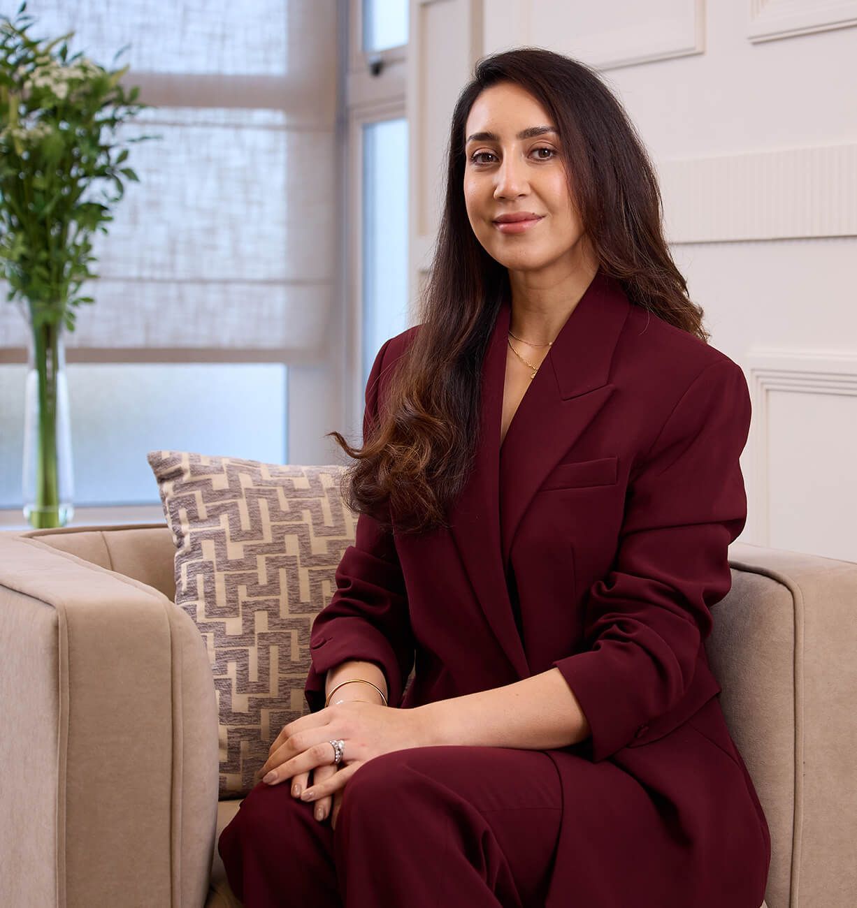 Woman in a burgundy suit seated on a beige sofa in a bright living room, smiling at the camera.
