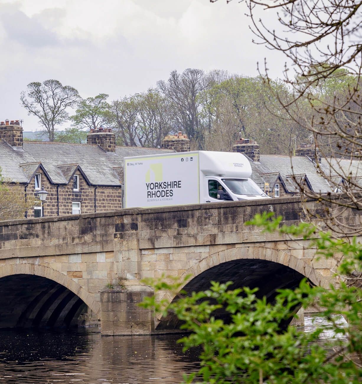 White delivery truck crosses an old stone bridge over a river, with stone buildings and trees behind.