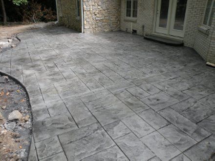 A gray, rectangular-patterned stamped concrete patio adjacent to a light-colored brick house.