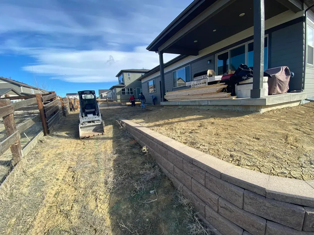 A small excavator parked on a dirt path next to a newly built home with a retaining wall on a sunny day.