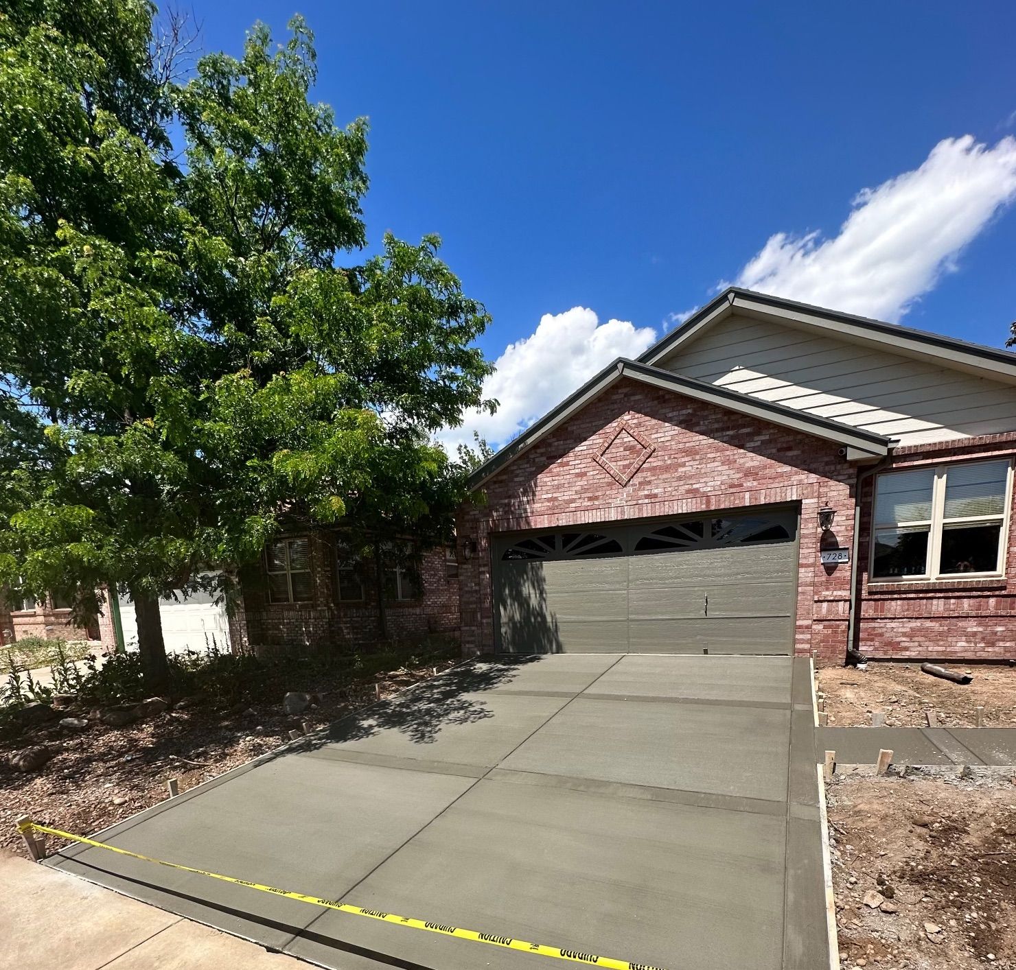 Brick suburban house with closed double garage, sloped driveway, with concrete slab in Longmont and a tree in the front yard under a blue sky