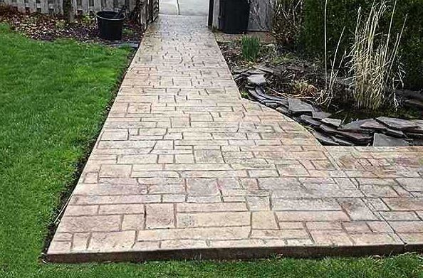 A walkway made of stamped concrete in a rectangular pattern, flanked by green grass on the left and a stone garden bed.