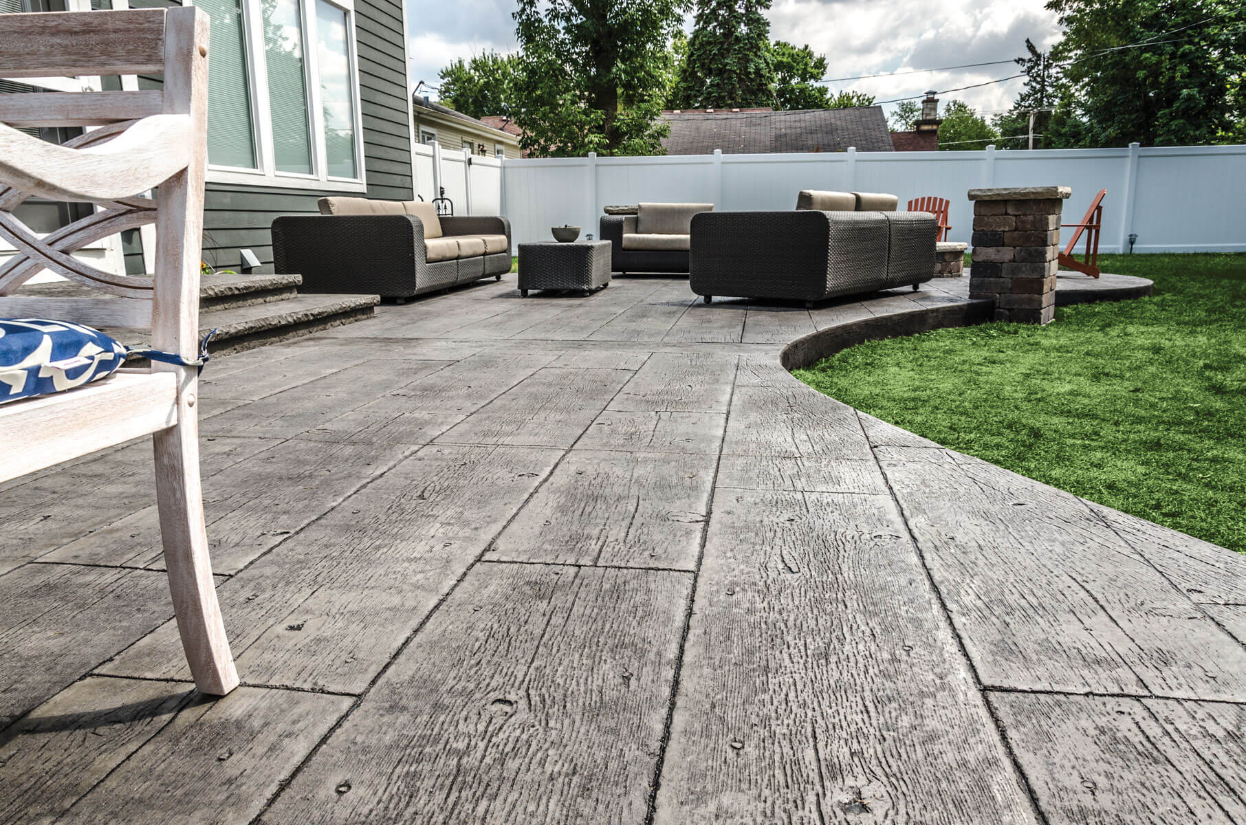 A patio with grey wood-plank stamped concrete, outdoor furniture, and a grassy yard against a white vinyl fence.