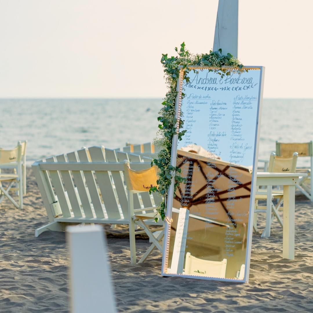 Tabella dei posti a sedere per matrimoni sulla spiaggia con specchio decorato e ghirlanda floreale.