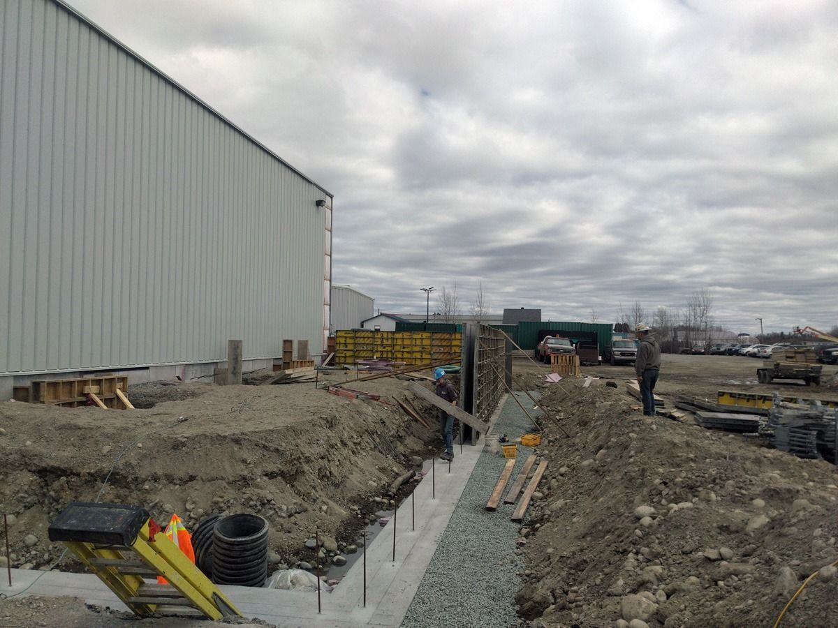 Chantier de construction avec une longue tranchée jouxtant un bâtiment métallique, ciel couvert.