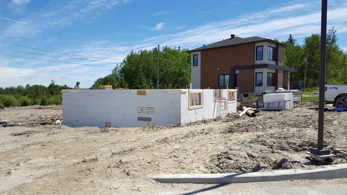 Fondations d'une maison en construction à côté d'une maison partiellement construite, ciel bleu en arrière-plan.