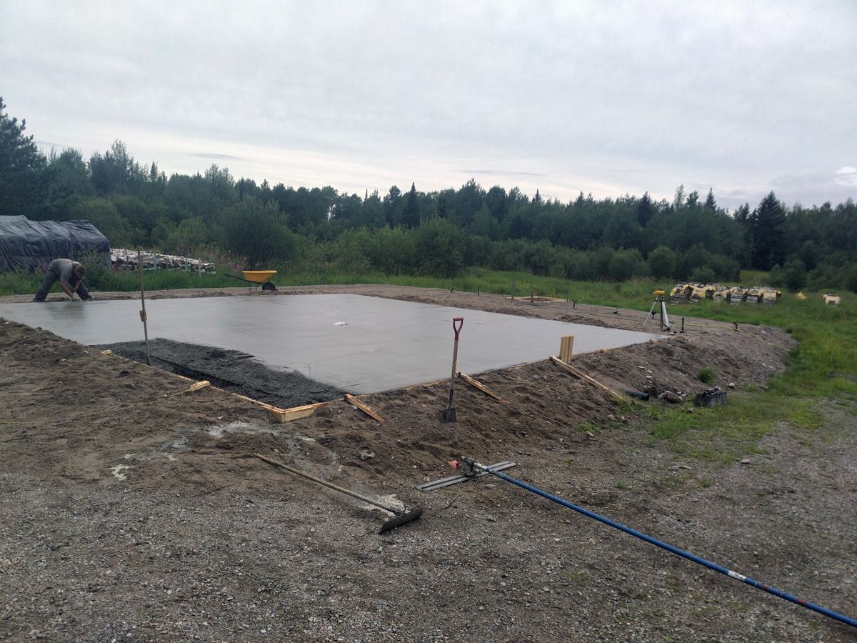 Coulage d'une fondation en béton, entourée de terre et de gravier, avec des arbres en arrière-plan.