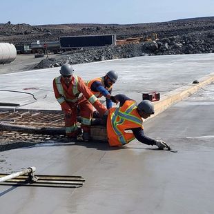 Trois ouvriers du bâtiment lissent du béton frais sur une grande dalle ; deux portent des vêtements orange, le troisième un gilet de sécurité.