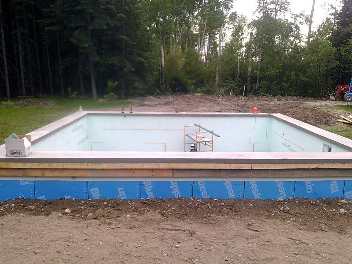 Piscine en construction : coffrage rectangulaire en béton avec isolation bleue, entourée de terre et d'arbres.