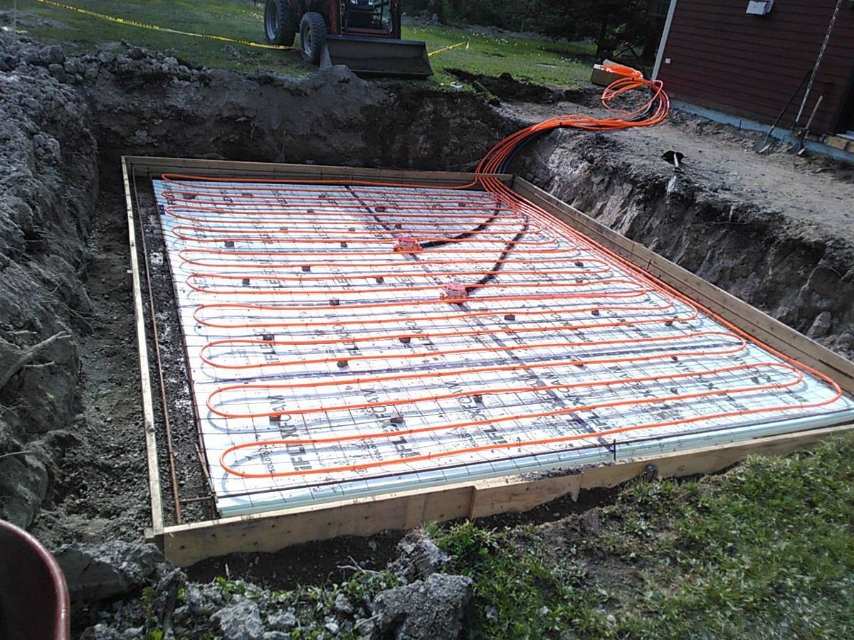 Fosse de construction encadrée avec tubes de chauffage radiant rouges posés sur une isolation blanche, prête pour le coulage du béton.