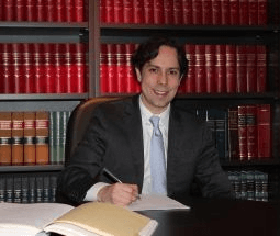 A man in a suit and tie is sitting at a desk in front of a bookshelf.