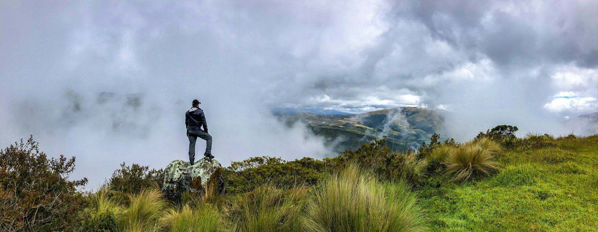A man is standing on top of a grassy hill looking at the clouds.