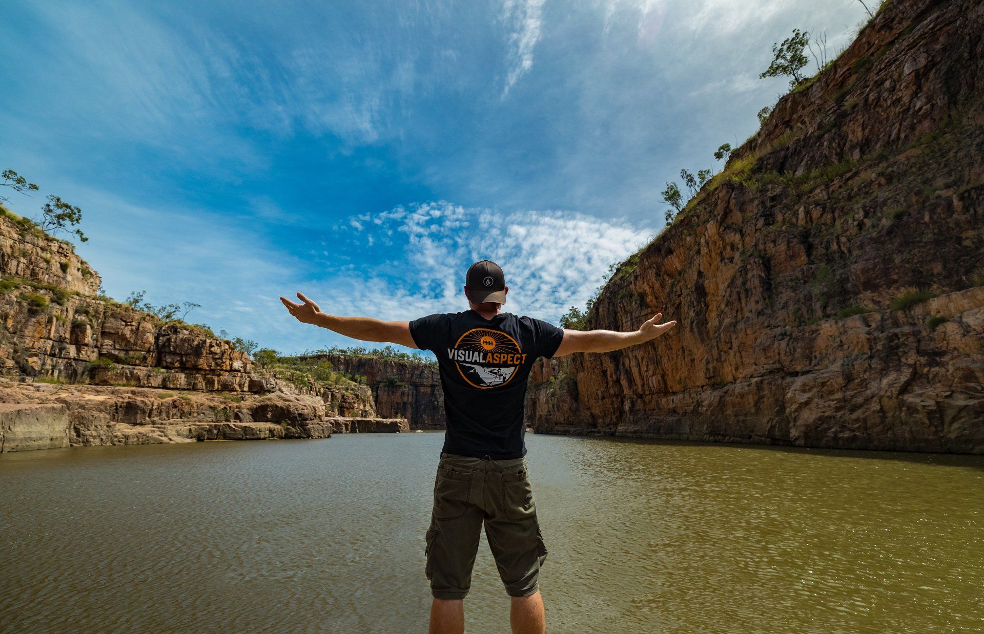 A man is standing in front of a lake with his arms outstretched.