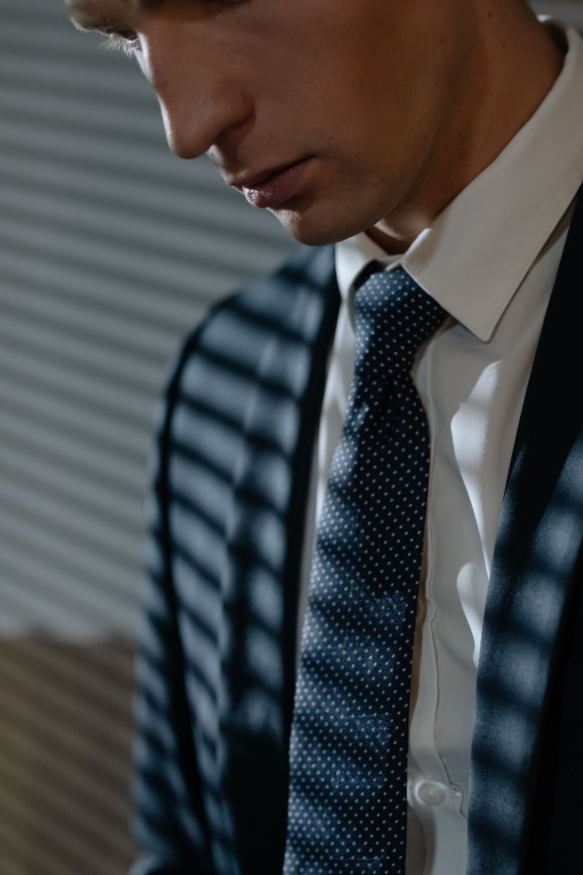 Man in a suit and tie lit by striped window shadows, looking down in a dim office setting