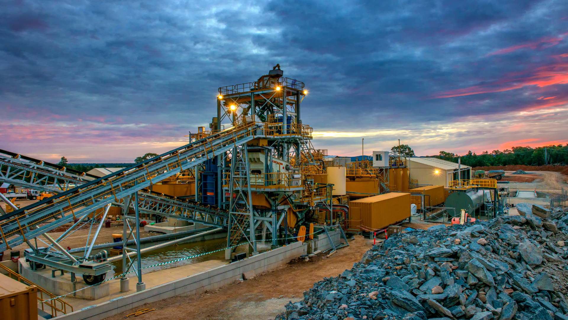 Complejo minero industrial al anochecer con cintas transportadoras, maquinaria y montones de rocas bajo un cielo colorido.
