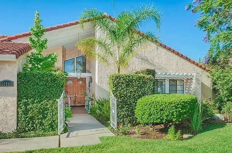 Beige house with red tile roof, arched entryway, palm tree, and well-manicured hedges.