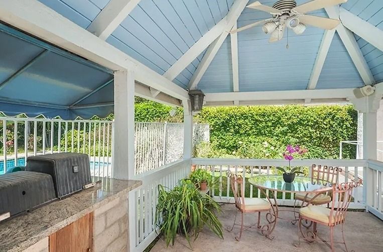 Covered patio with grill, table, chairs, and pool view, blue ceiling, white railing, greenery.