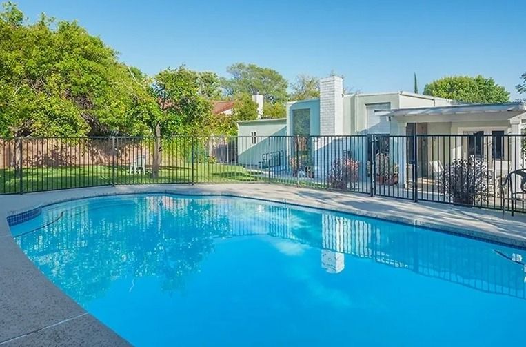 Swimming pool surrounded by a black fence and greenery, with a light-colored house in the background under a blue sky.