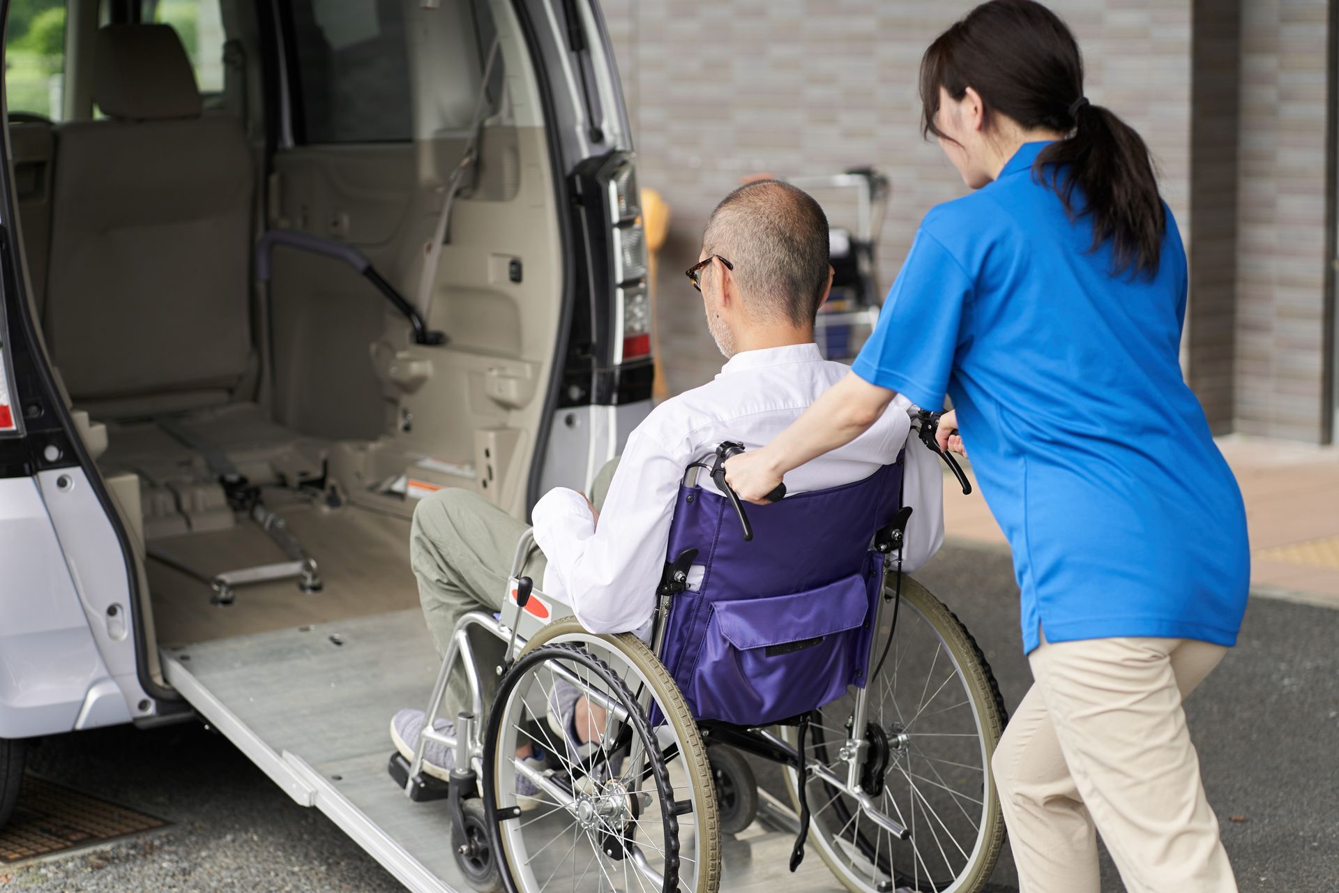 Woman assisting person in wheelchair onto accessible van with a ramp.