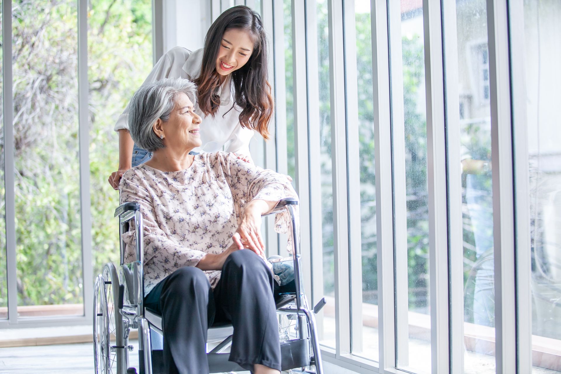 Woman in wheelchair looks out window with caregiver smiling beside her.