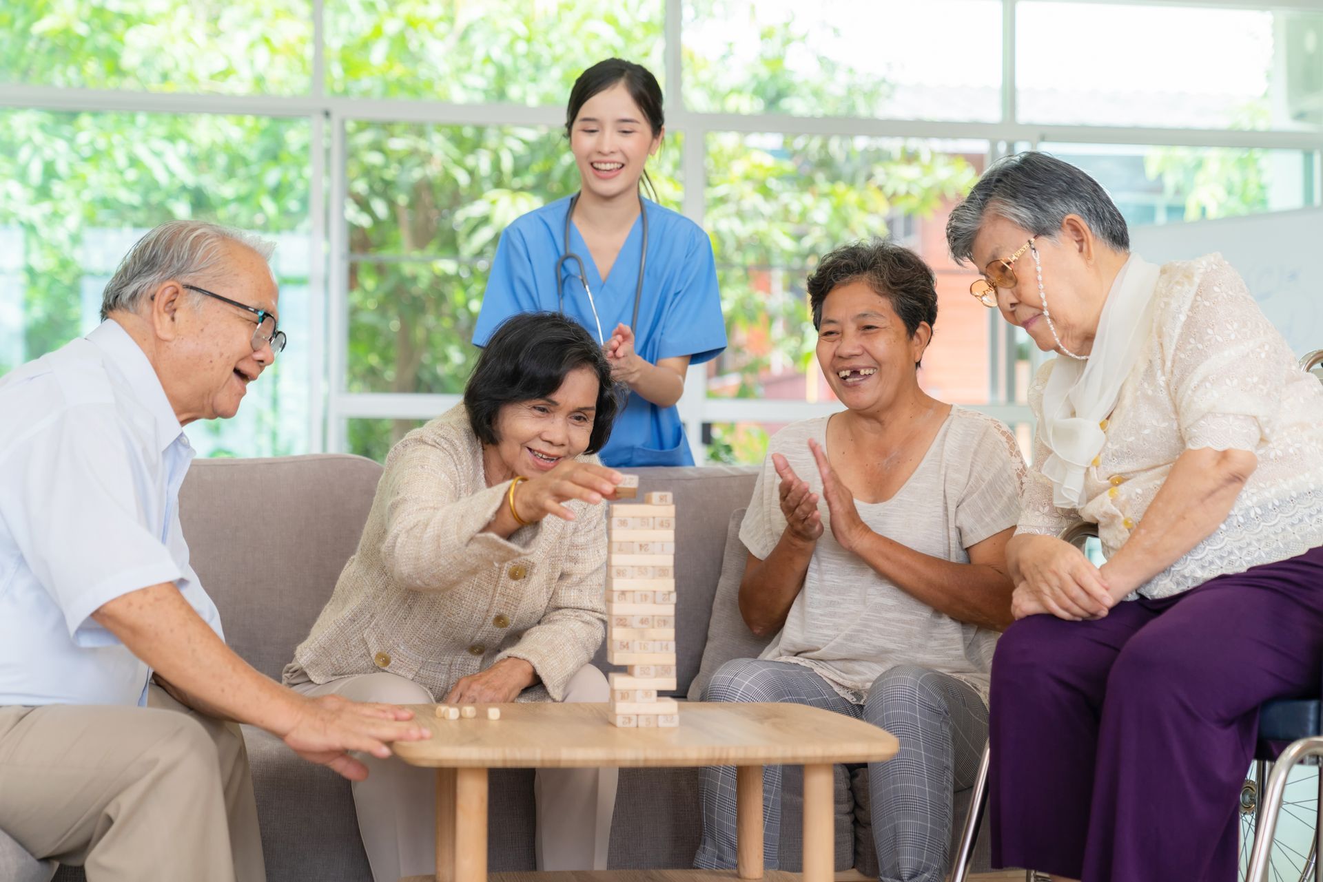 Seniors playing Jenga with a healthcare worker. People smile and clap in a living room with large windows.