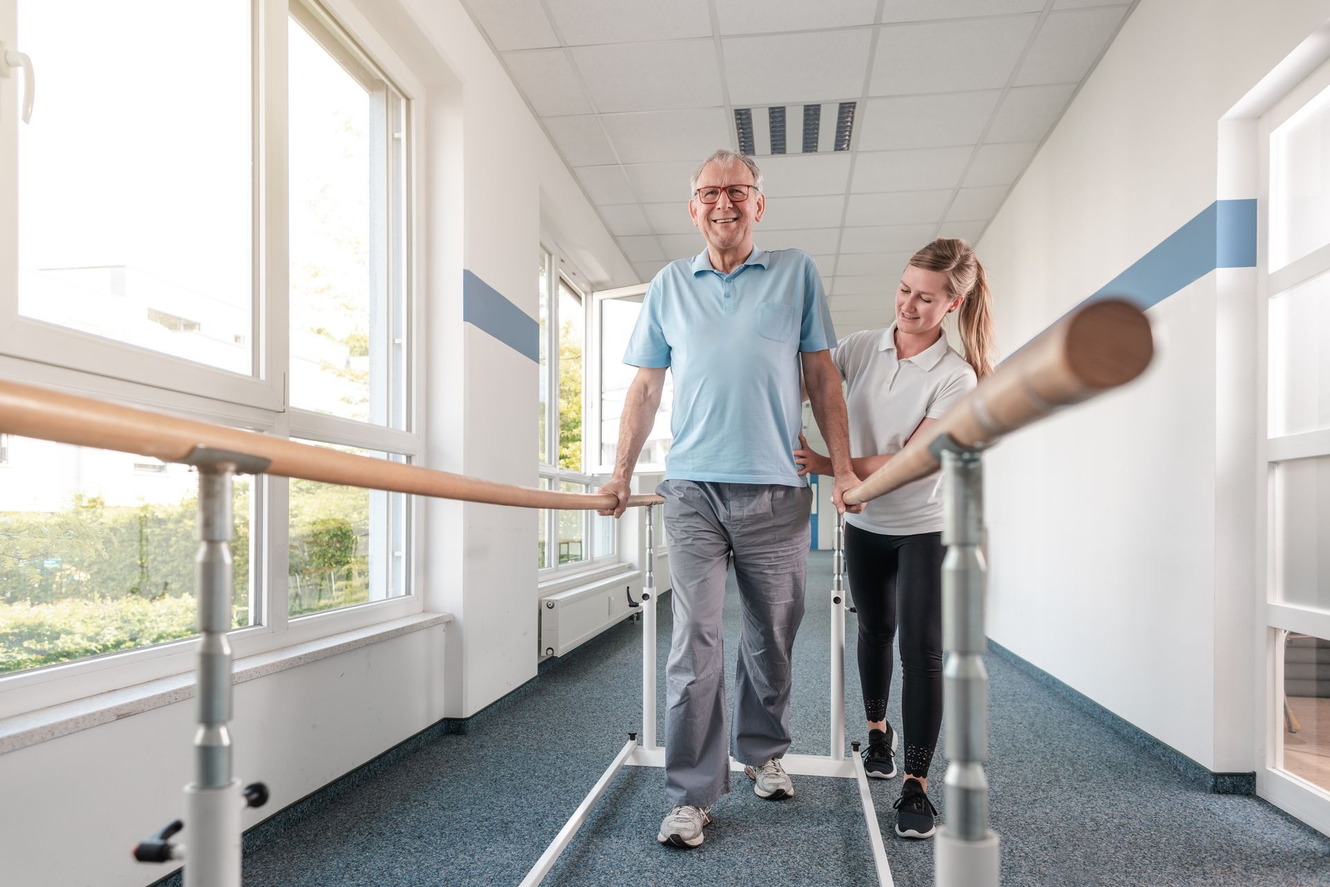 Man walking with a therapist in a rehabilitation hallway, using parallel bars.