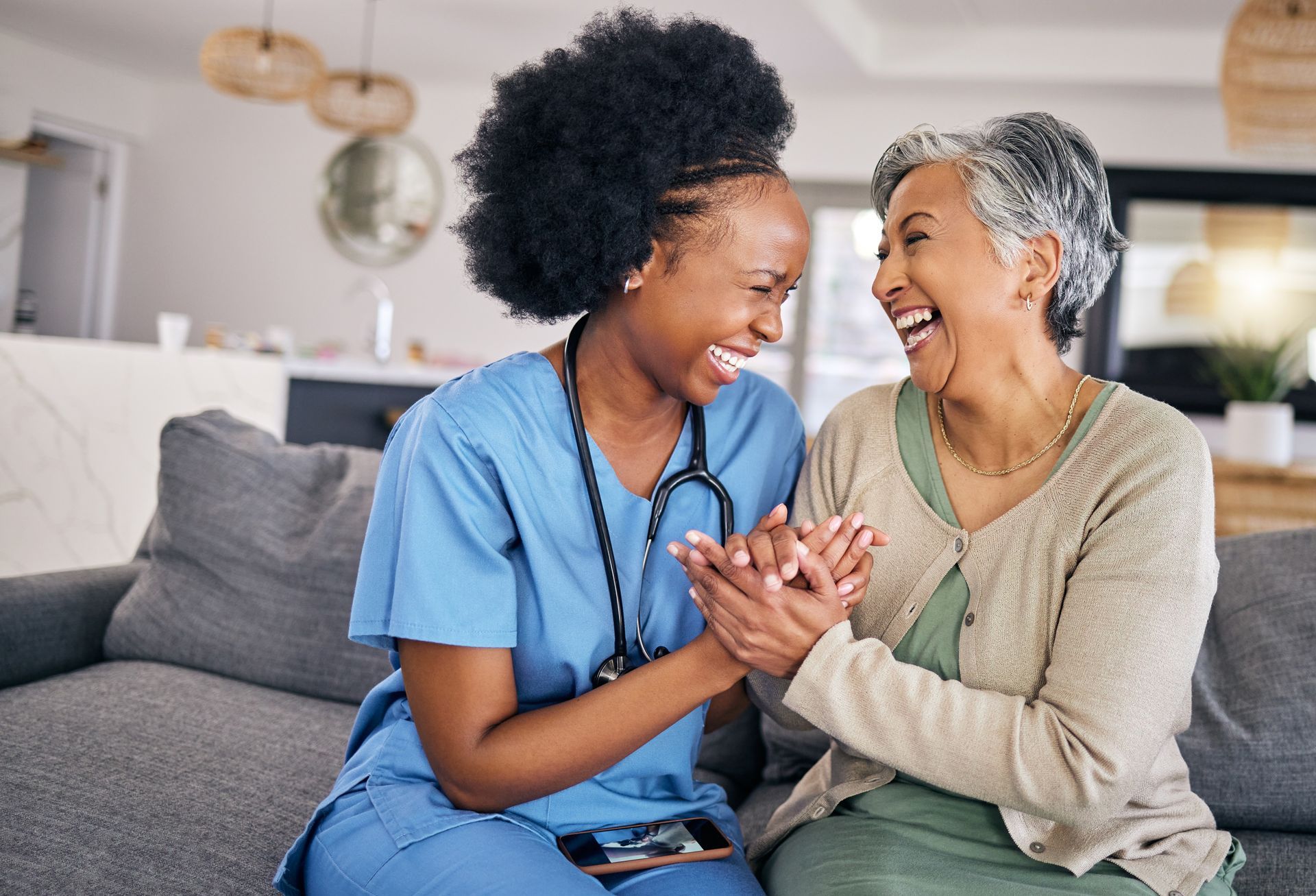 Smiling caregiver in blue scrubs holding hands with laughing senior in a home setting.