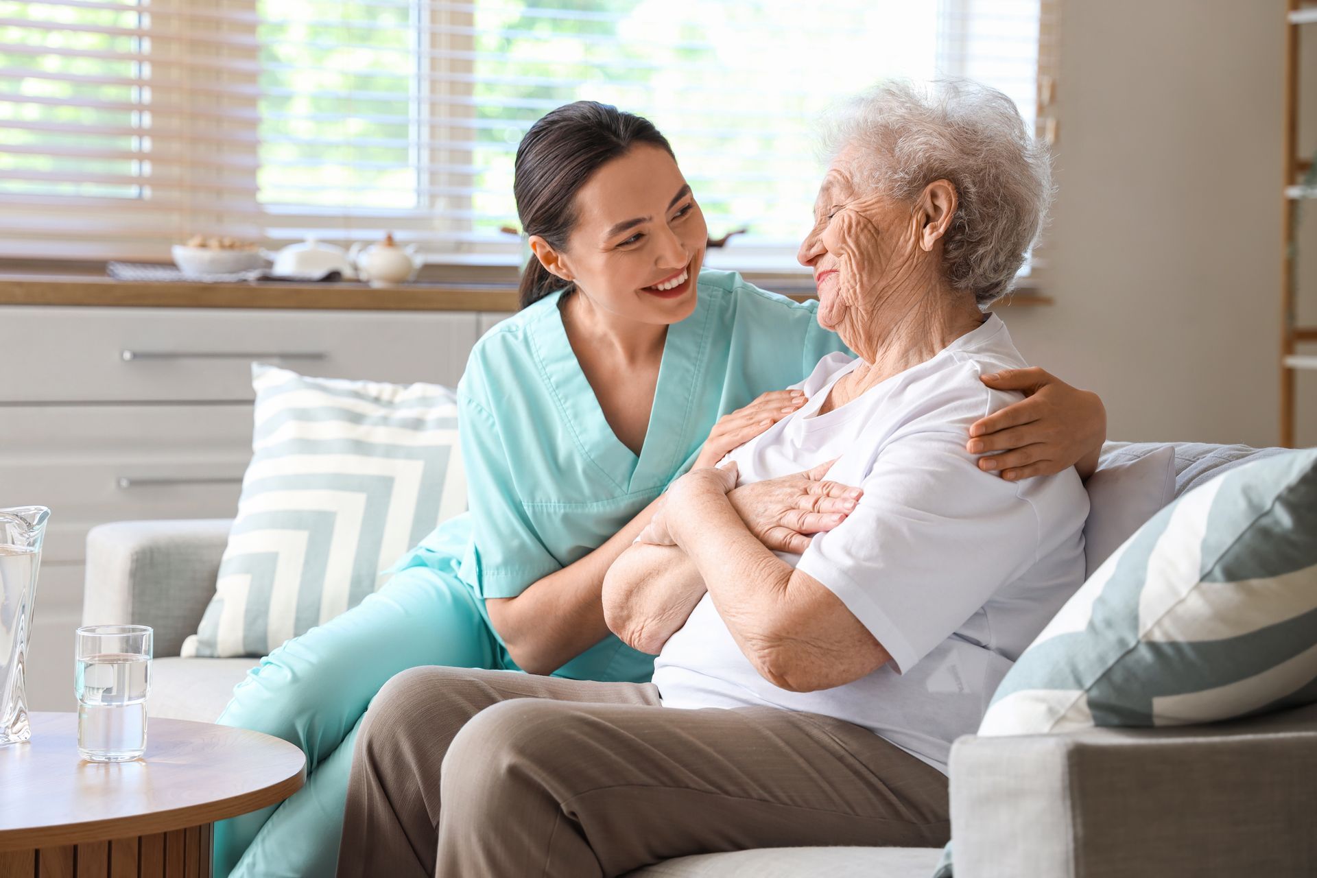 Caregiver in teal scrubs comforts an elder woman on a couch; warm interior, smiles, and touch.