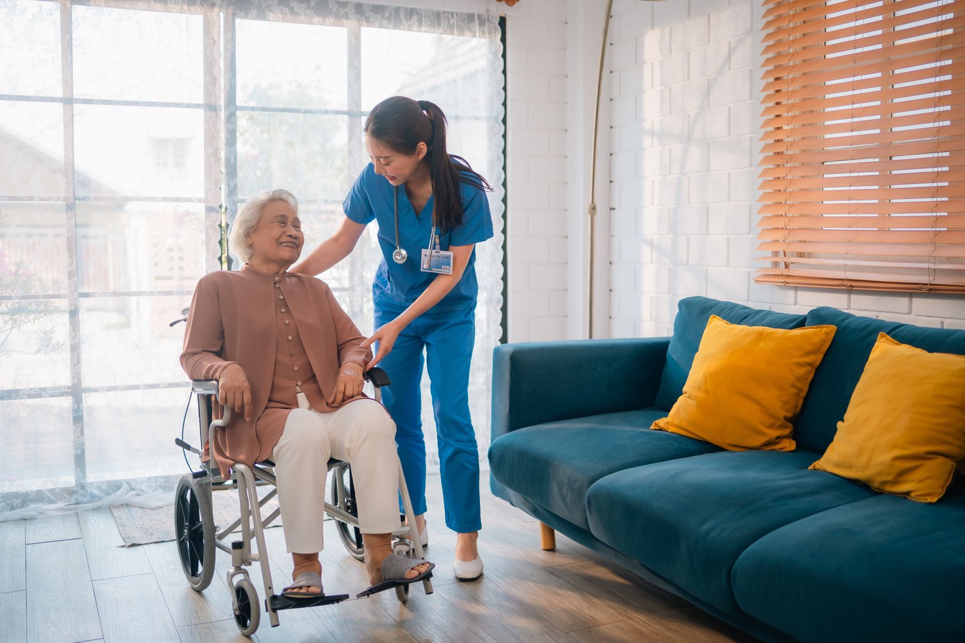 A caregiver in blue scrubs assists an older person in a wheelchair indoors.