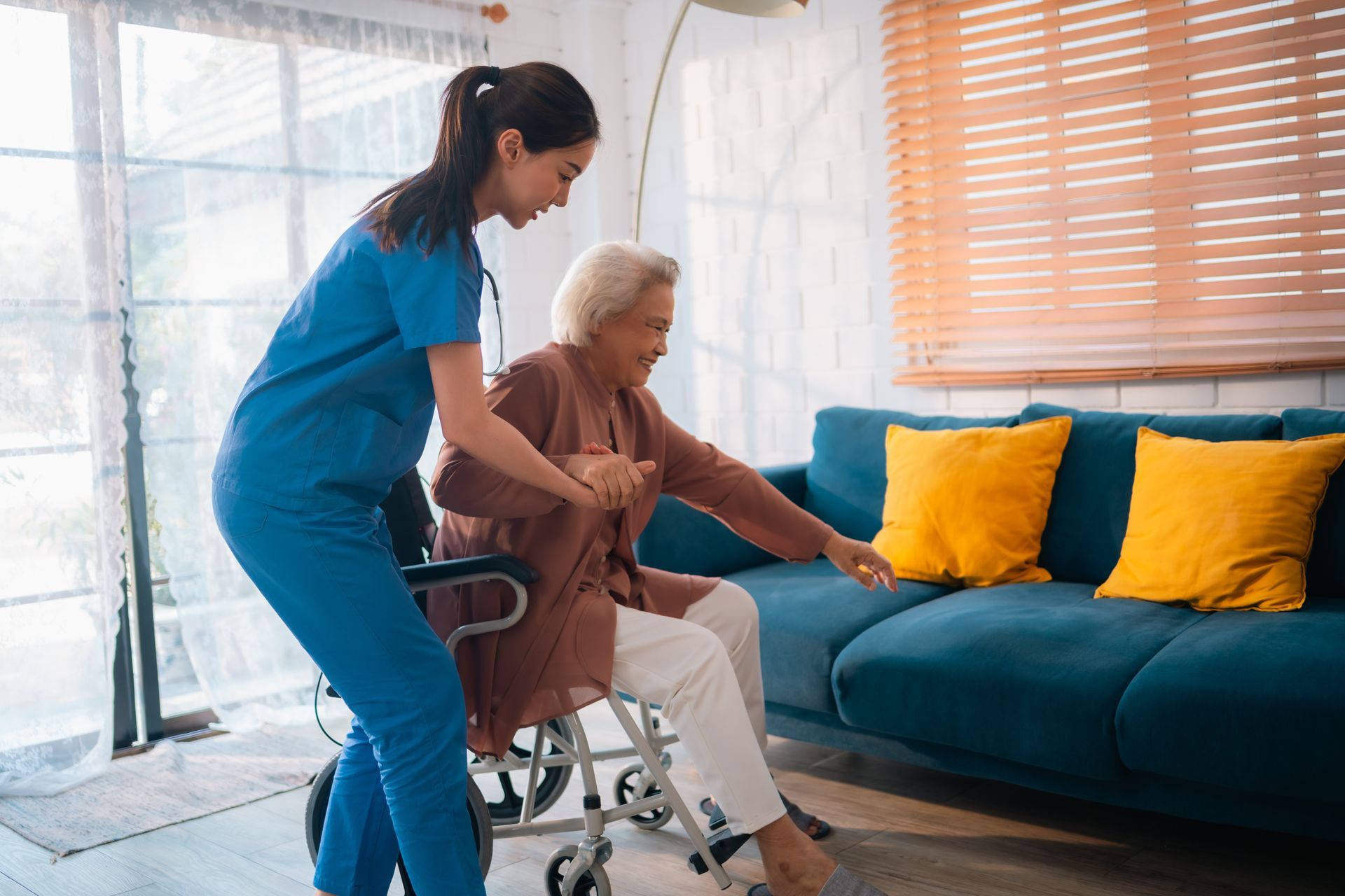 Caregiver assists an older person from a wheelchair to a sofa in a home.