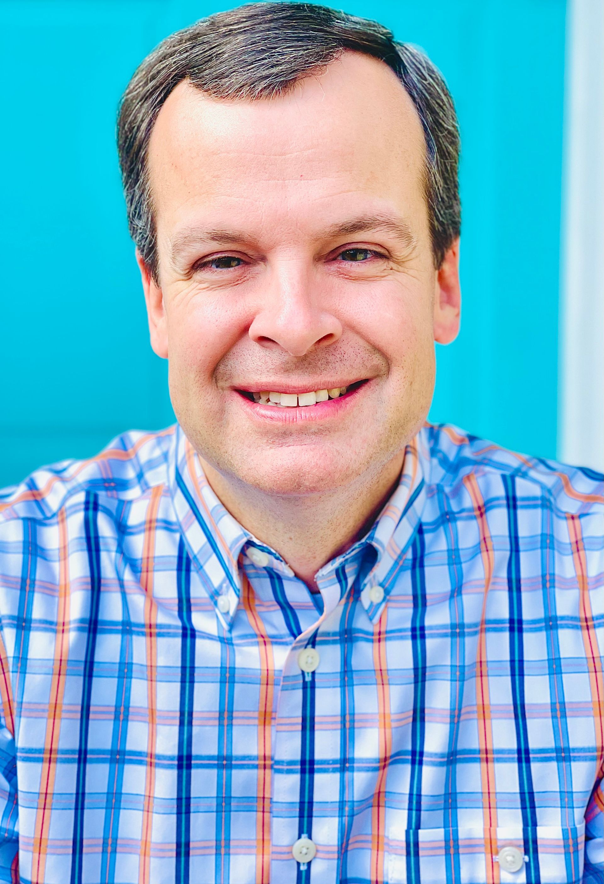 A man in a plaid shirt is smiling in front of a blue wall.