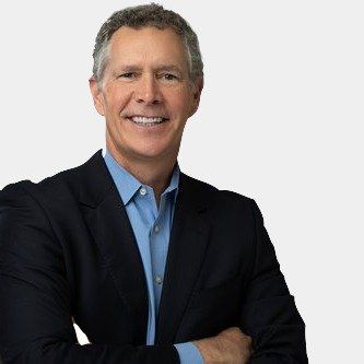 A man in a suit and blue shirt is smiling with his arms crossed.