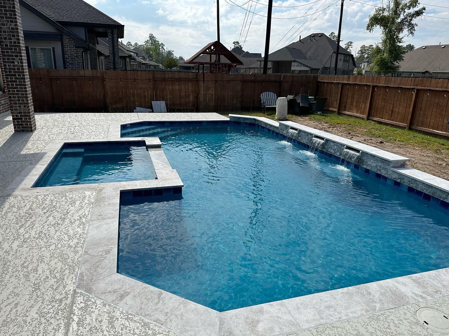 Backyard swimming pool with attached hot tub, blue water, stone deck, and wooden fence
