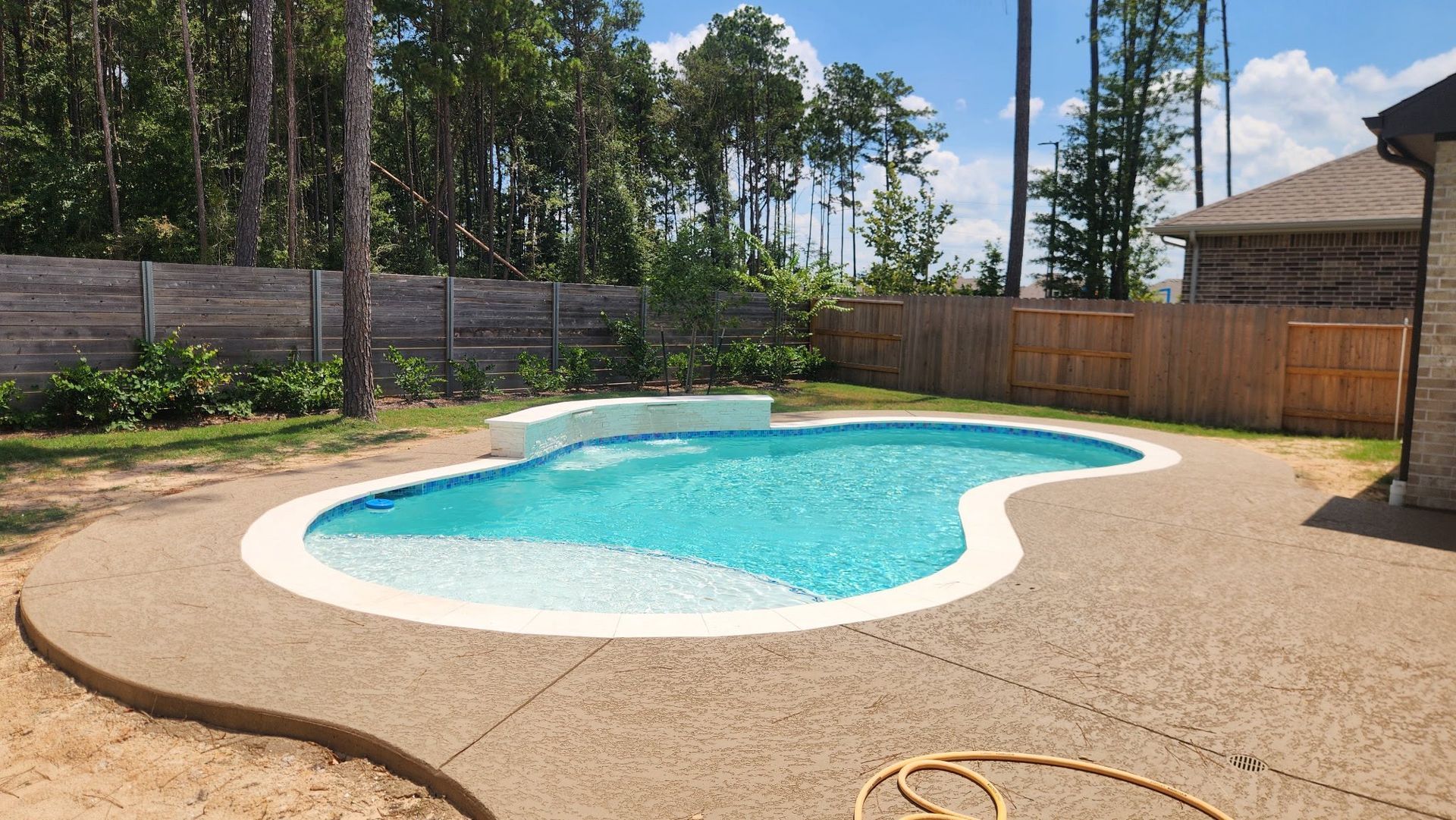 Backyard swimming pool with bright blue water, tan deck, wooden fence, and trees under a sunny sky