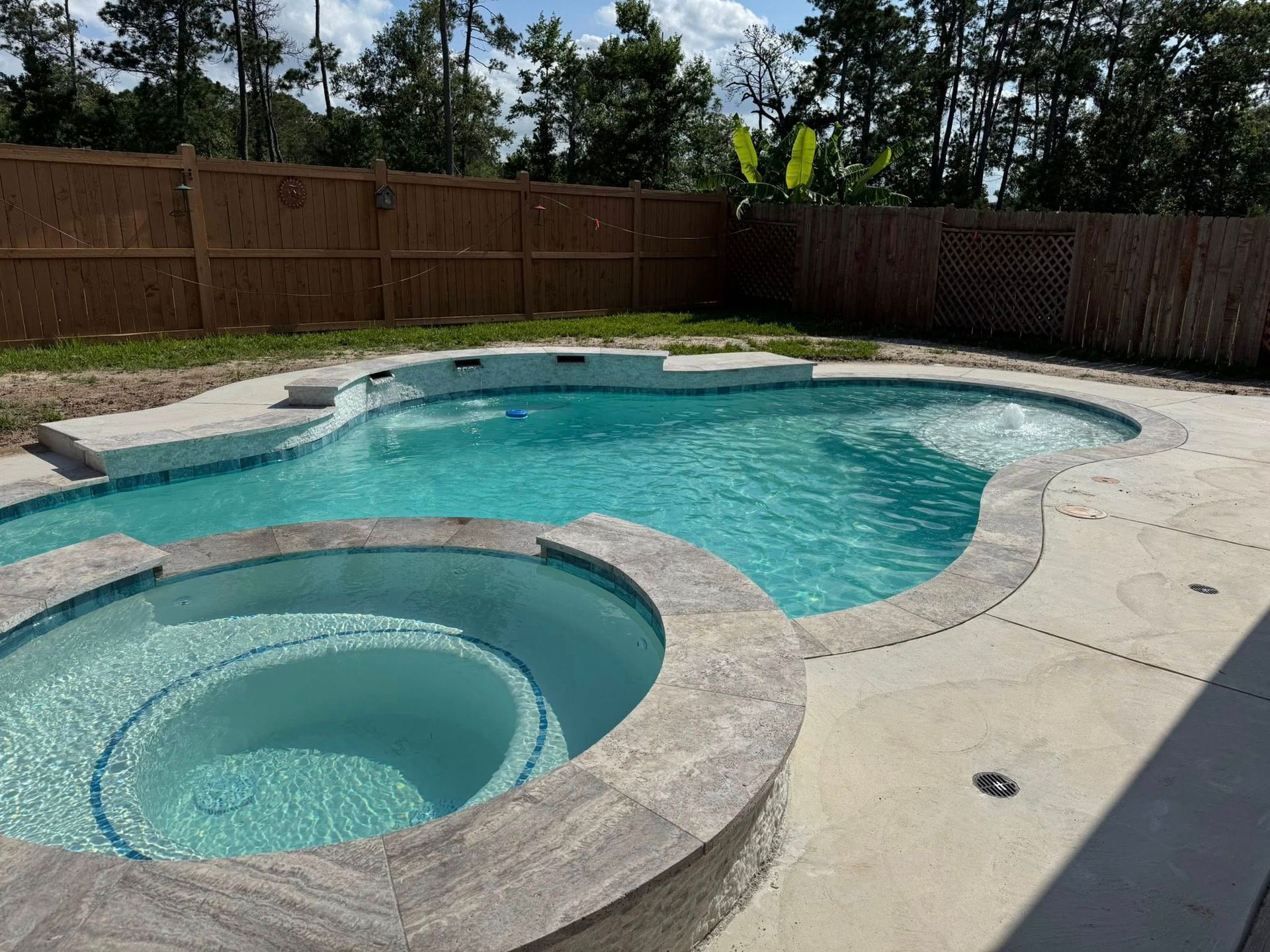 Backyard swimming pool with a hot tub and wooden privacy fence in bright sunlight