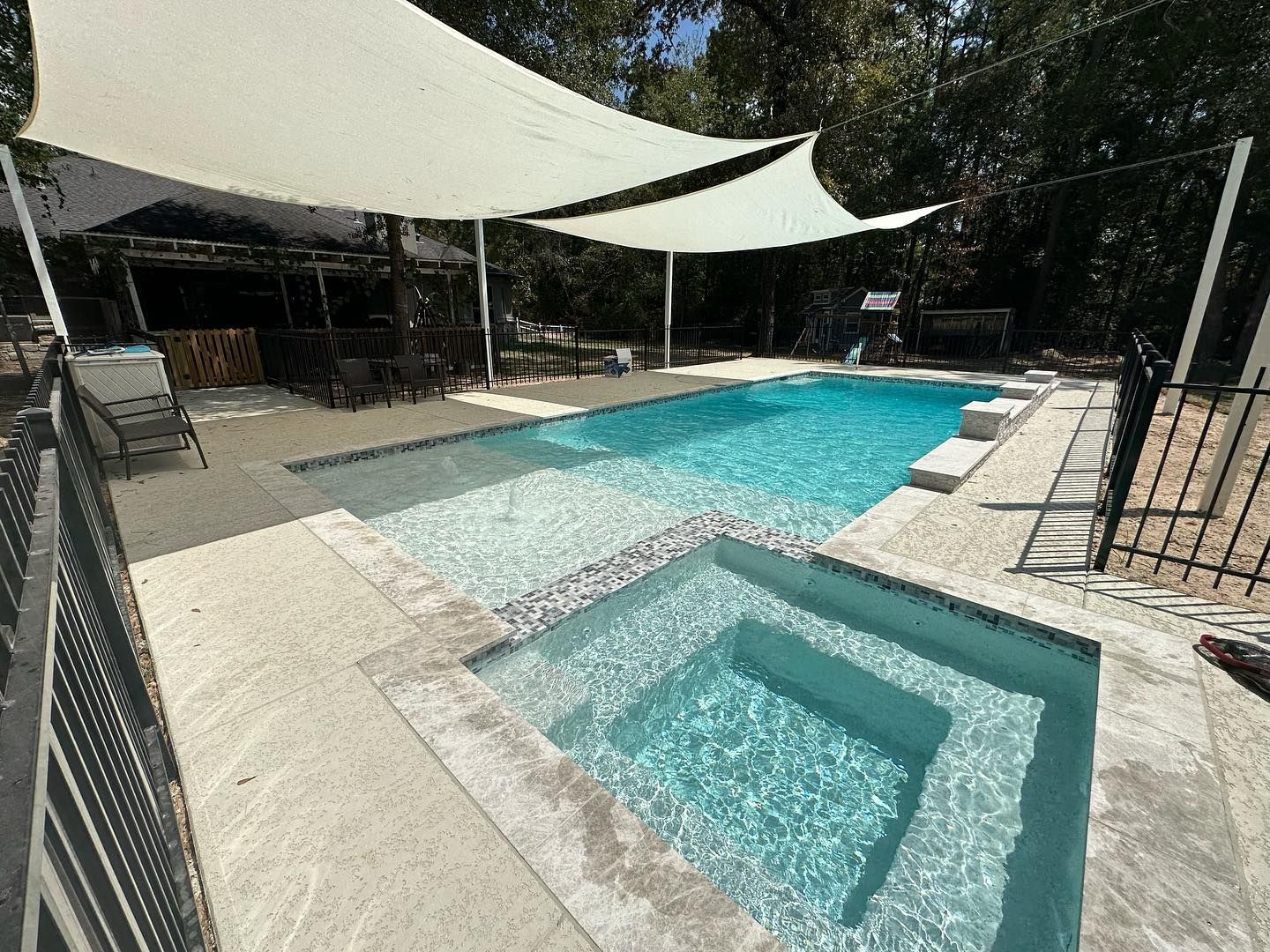 Backyard pool with a hot tub, white shade sails, and lounge chairs in bright sunlight