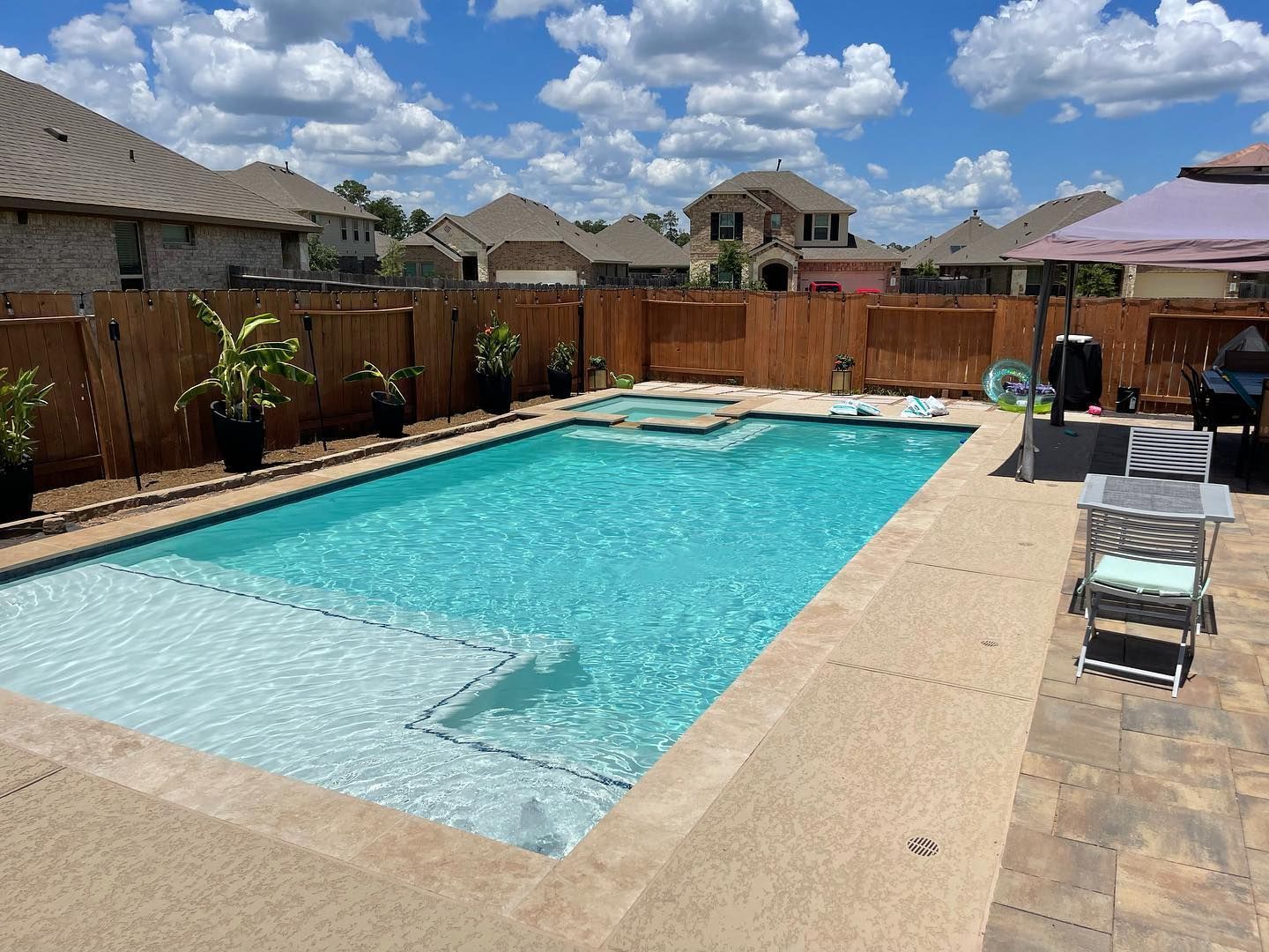 Backyard swimming pool with turquoise water, lounge chairs, wooden fence, and houses under a cloudy blue sky