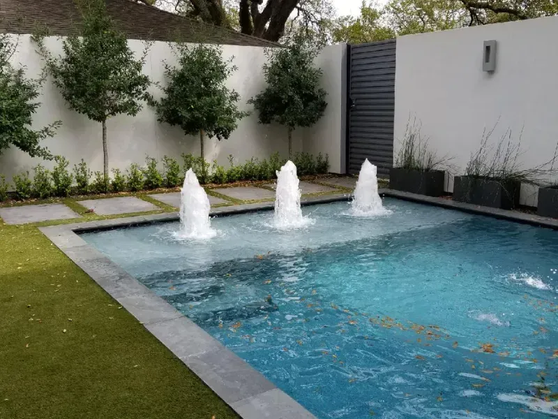 Modern backyard pool with three water fountains, blue water, and surrounding greenery