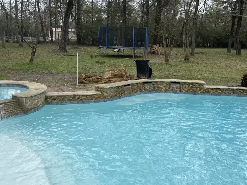 Backyard swimming pool with stone edging and a trampoline in the grassy yard beyond.