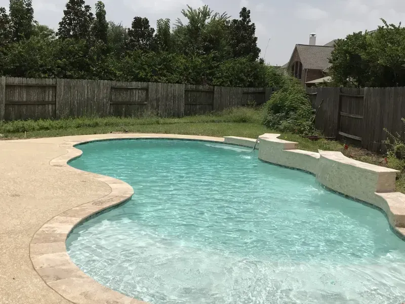 Backyard swimming pool with clear water, stone edge, fence, and trees in the background