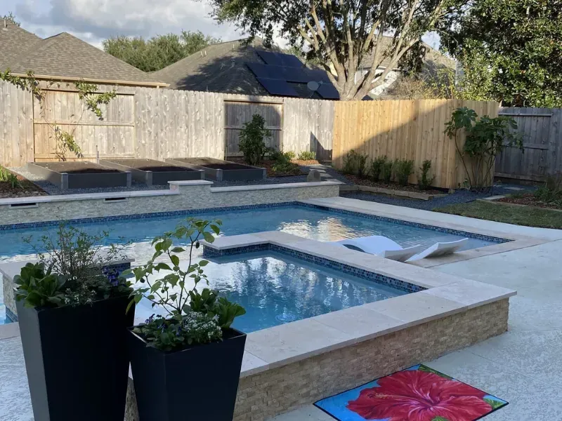 Backyard pool with spa, tan pavers, wooden fence, and potted plants in the foreground