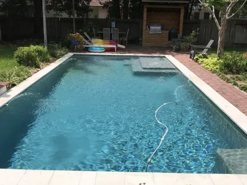 Backyard swimming pool with blue water and a hose floating in it, bordered by brick and greenery