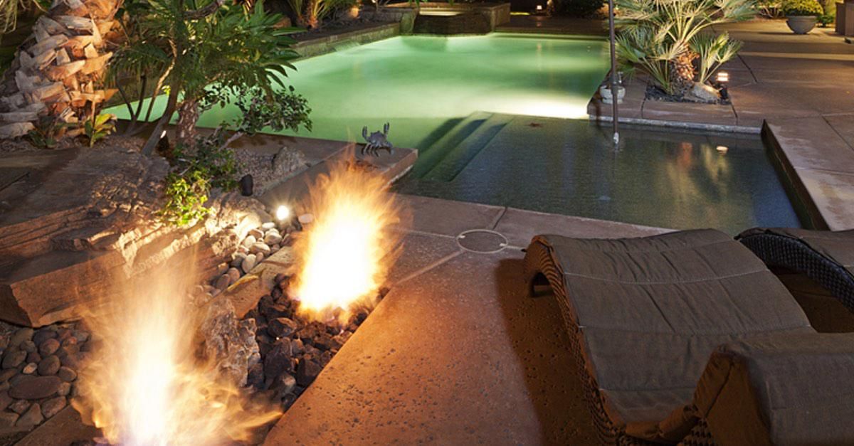 Nighttime poolside patio with a lit fire pit and lounge chairs beside a lit swimming pool